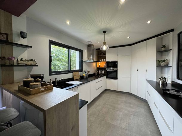 Cuisine contemporaine spacieuse avec des façades blanches, un plan de travail noir, des étagères ouvertes et un îlot en bois, éclairée naturellement par une grande fenêtre avec vue sur l'extérieur.