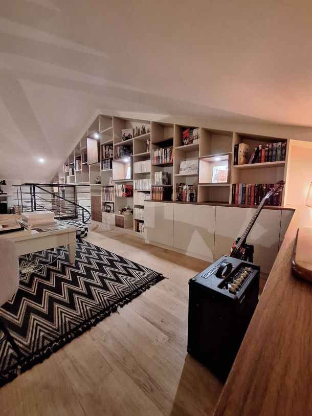 Aménagement de bibliothèque sur mesure sous pente avec étagères ouvertes et rangements fermés, intégrée dans un espace chaleureux avec un sol en parquet clair, un tapis graphique noir et blanc, et des éléments décoratifs comme une guitare et un ampli.